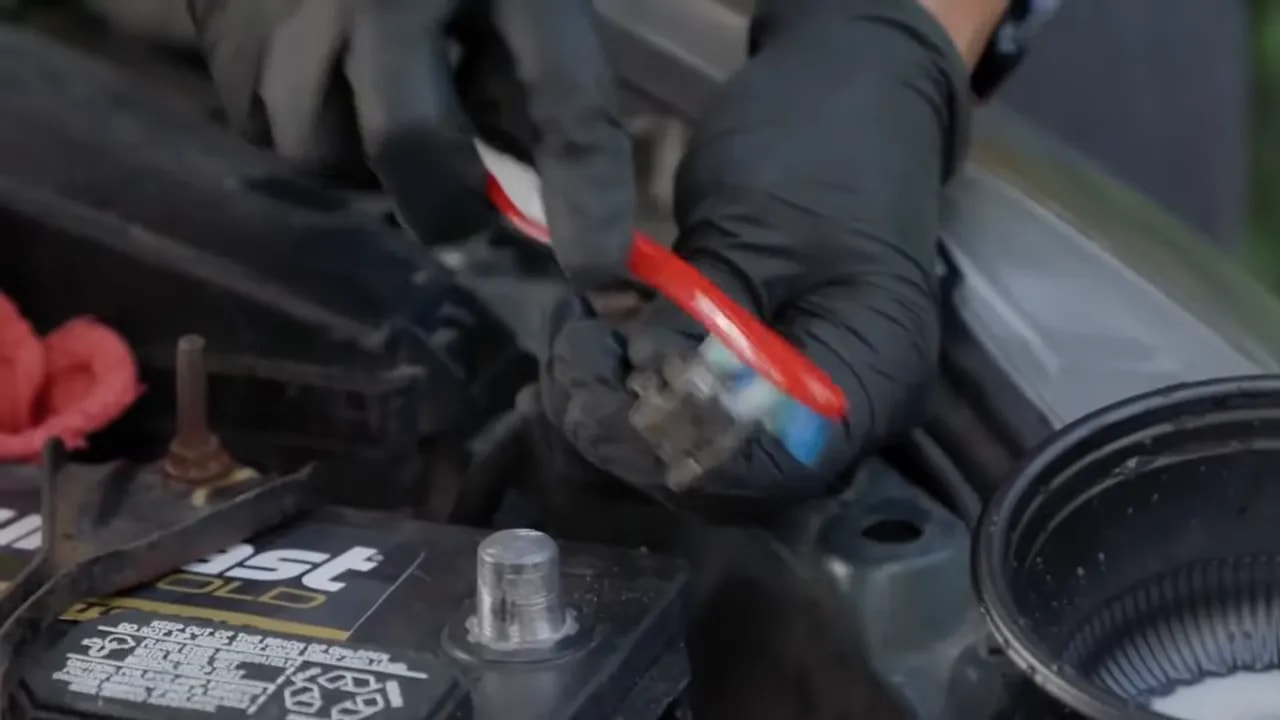Gloved hands brushing a disconnected battery cable clamp beside exposed battery posts