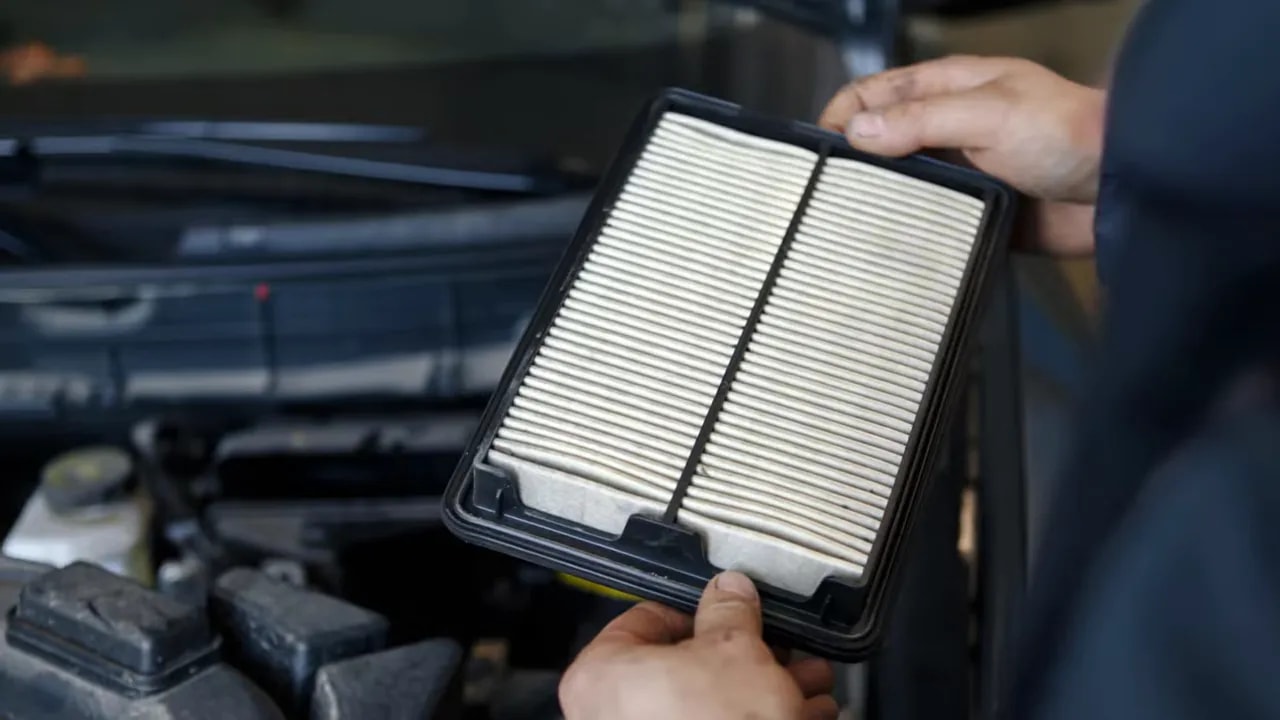 Hands holding a gray pleated air filter above an engine bay with a white filter nearby