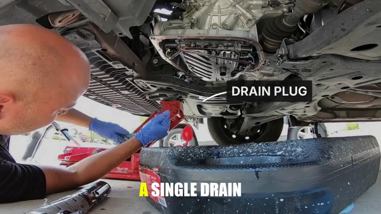 Technician beneath a raised vehicle with a label pointing to the transmission drain plug and a catch pan below