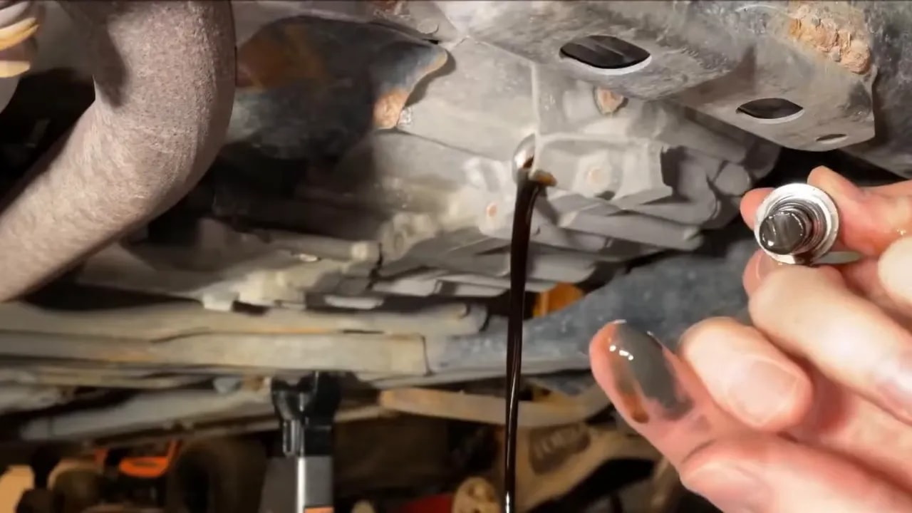 Dark fluid draining beneath a vehicle beside a hand holding the drain plug